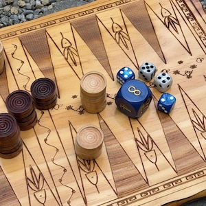 May include: A wooden backgammon board featuring a natural grain finish with dark brown and light tan triangular points. The board includes stacks of light and dark wooden checkers, two white dice, two blue dice, and one large blue doubling cube.