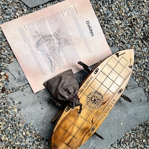 Puede incluir: Un juego de mesa Daldos de madera con un acabado marr&oacute;n claro, con l&iacute;neas de cuadr&iacute;cula y runas grabadas. Tambi&eacute;n hay una peque&ntilde;a bolsa con cord&oacute;n y un papel con el nombre y las instrucciones del juego. El juego est&aacute; sobre una superficie de piedra.