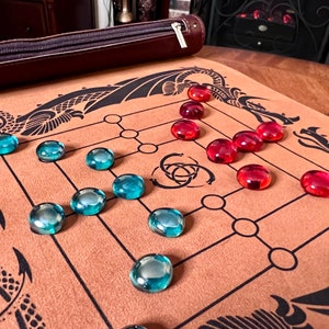 May include: A brown leather game board with black dragon designs and a Celtic knot symbol in the center. The board has a grid pattern with blue and red glass game pieces.