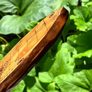 Puede incluir: Primer plano de un tablero de juego de madera con una superficie marr&oacute;n claro y bordes marrones oscuros. El tablero presenta l&iacute;neas grabadas y dise&ntilde;os circulares. El fondo es follaje verde borroso.