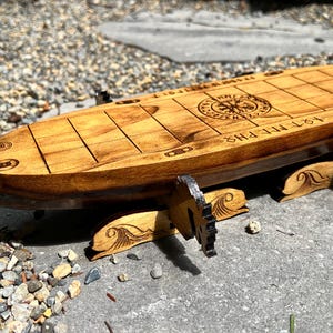 Puede incluir: Un tablero de juego de madera con forma de barco con un dise&ntilde;o de rosa de los vientos y texto grabado. El tablero es de madera de color marr&oacute;n claro y tiene un patr&oacute;n de cuadr&iacute;cula. Los soportes tienen un dise&ntilde;o de olas. El texto dice "1ST MEPHIS".
