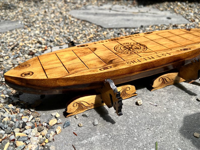 Puede incluir: Un tablero de juego de madera con forma de barco con un dise&ntilde;o de rosa de los vientos y texto grabado. El tablero es de madera de color marr&oacute;n claro y tiene un patr&oacute;n de cuadr&iacute;cula. Los soportes tienen un dise&ntilde;o de olas. El texto dice "1ST MEPHIS".
