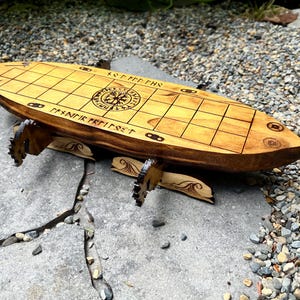 Puede incluir: Un juego de mesa vikingo de madera con un acabado marr&oacute;n claro. El tablero tiene un patr&oacute;n de cuadr&iacute;cula y runas y s&iacute;mbolos grabados. El tablero est&aacute; soportado por patas decorativas de madera. El juego est&aacute; sobre una superficie de piedra.