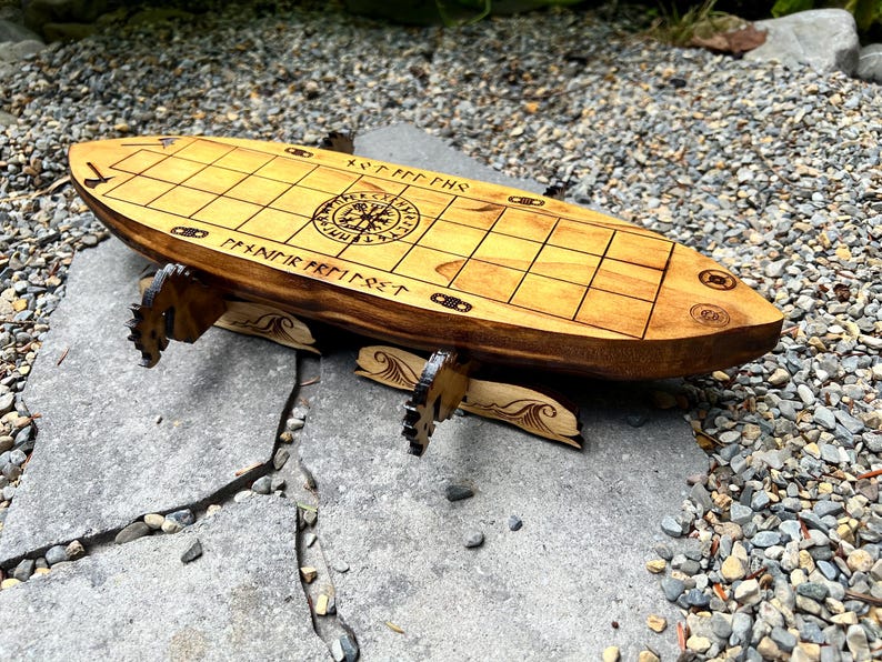 Puede incluir: Un juego de mesa vikingo de madera con un acabado marr&oacute;n claro. El tablero tiene un patr&oacute;n de cuadr&iacute;cula y runas y s&iacute;mbolos grabados. El tablero est&aacute; soportado por patas decorativas de madera. El juego est&aacute; sobre una superficie de piedra.