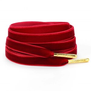 Puede incluir: Cordones de terciopelo rojo con aglets dorados. Los cordones están doblados y dispuestos en círculo. Los cordones tienen el texto "LOOP KING" en los aglets.