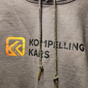 Op de afbeelding: Een grijze hoodie met een zwart logo dat "Kompelling Kars" zegt en een gele en oranje grafische afbeelding van een gestileerde pijl. De hoodie heeft een trekkoord met gouden uiteinden.
