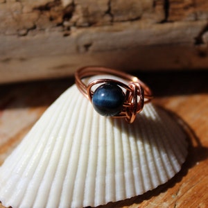 May include: A copper wire wrapped ring with a blue tiger's eye gemstone. The ring is sitting on a white seashell.