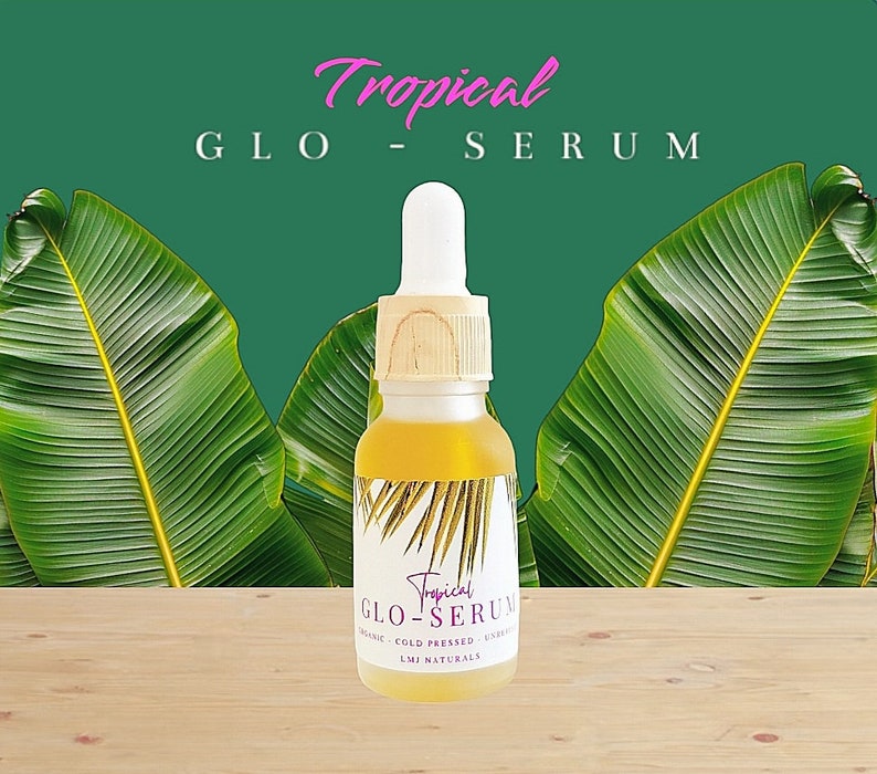 May include: A glass bottle of Tropical Glo Serum with a wooden cap. The bottle is sitting on a wooden surface with green palm leaves in the background. The label on the bottle says "Tropical Glo Serum" and "Organic Cold Pressed - Unrefined - LMI Naturals".