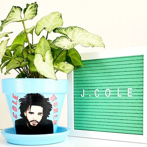 May include: A light blue ceramic planter with a graphic of J. Cole's face and the words "J. Cole World" on the front. The planter is decorated with a green and white leafy plant.