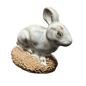 May include: A white ceramic rabbit figurine sitting on a brown ceramic base. The rabbit has long ears and a detailed face.