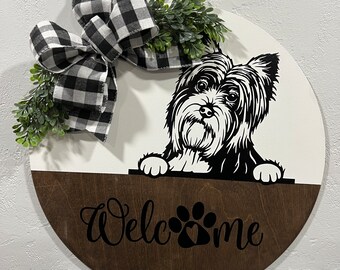 May include: A round wooden welcome sign featuring a black and white dog illustration. The sign has a white top half and a brown bottom half with the word "Welcome" and paw prints. A black and white checkered bow and greenery are attached.