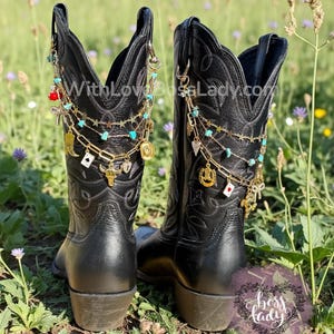 May include: Two black cowboy boots adorned with gold and turquoise boot chains. The chains feature charms including playing cards and stars. The boots are set in a field of green grass and wildflowers.
