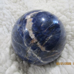 May include: A polished, spherical sodalite gemstone. The stone is a deep blue color with white and light gray veining throughout. The surface is smooth and reflective, with a slight sheen. The gemstone is set against a white, textured background.