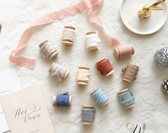 May include: Assortment of 12 spools of silk ribbon in various colors, including dusty rose, beige, light blue, and gray. The spools are made of wood and have a natural finish. The ribbon is arranged on a white linen background. The text "Her Vows" is visible on a small notebook in the lower left corner of the image.