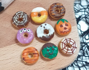 May include: Nine Halloween-themed mini donuts with various designs, including a spiderweb, a ghost, a pumpkin, and a candy corn. The donuts are arranged on a wooden surface.