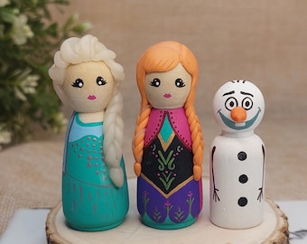 Può includere: Tre bambole di legno dipinte a mano, due ragazze e un pupazzo di neve. Le ragazze sono vestite come Elsa e Anna del film Frozen. Elsa indossa un vestito blu e argento con una treccia. Anna indossa un vestito viola e nero con una treccia rossa e arancione. Il pupazzo di neve indossa un cappello bianco e ha un naso di carota nero.