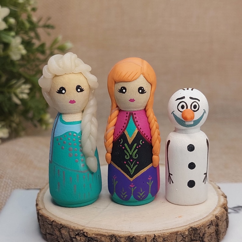 May include: Three hand-painted wooden dolls, two girls and a snowman. The girls are dressed as Elsa and Anna from the film Frozen. Elsa is wearing a blue and silver dress with a braid. Anna is wearing a purple and black dress with a red and orange braid. The snowman is wearing a white hat and has a black carrot nose.