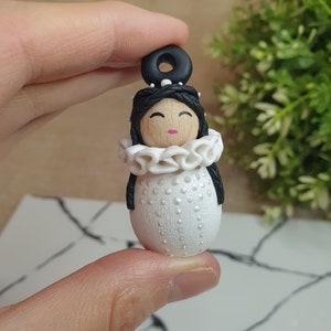 May include: A white polymer clay doll with black hair, a white ruffle collar, and a black headband. The doll has a wooden face with painted eyes, a pink mouth, and white dots on the body.