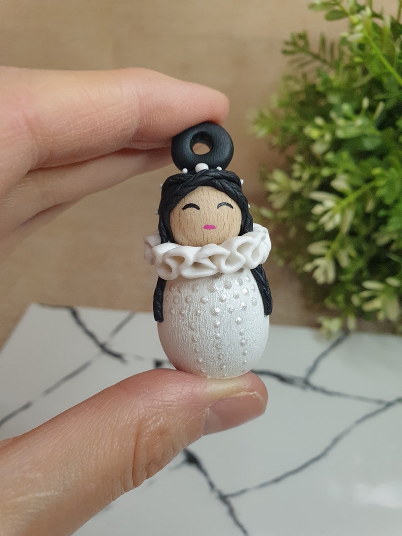 May include: A white polymer clay doll with black hair, a white ruffle collar, and a black headband. The doll has a wooden face with painted eyes, a pink mouth, and white dots on the body.