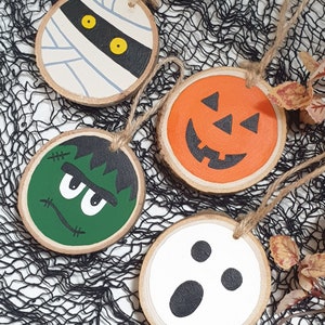 May include: Four wooden Halloween ornaments with painted faces. The ornaments are round and have a string attached for hanging. The faces are of a mummy, a pumpkin, Frankenstein, and a ghost.