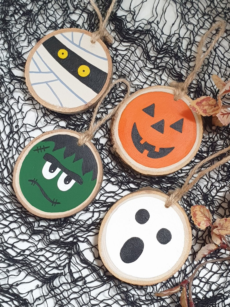 May include: Four wooden Halloween ornaments with painted faces. The ornaments are round and have a string attached for hanging. The faces are of a mummy, a pumpkin, Frankenstein, and a ghost.