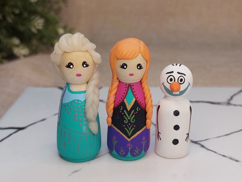 May include: Three hand-painted wooden peg dolls depicting characters from the movie Frozen. The dolls are Elsa with a light blue dress and blonde hair, Anna with a pink and purple dress and red hair, and Olaf the snowman with a carrot nose and black eyes.