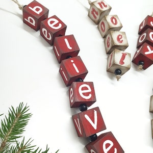 May include: Red wooden blocks with white letters spelling out "believe" hanging from a twine string. The blocks are strung together with black beads.