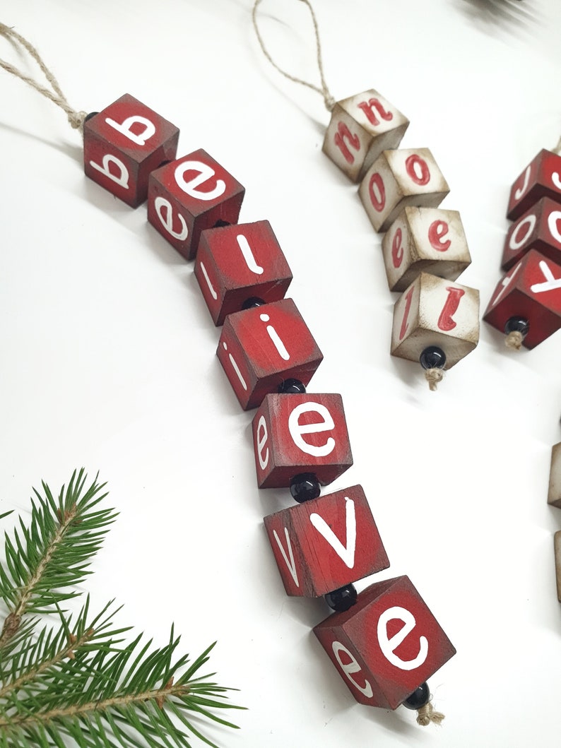 May include: Red wooden blocks with white letters spelling out "believe" hanging from a twine string. The blocks are strung together with black beads.