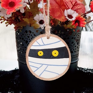 May include: A white and blue mummy face painted on a round wooden slice with yellow eyes. The slice is hanging from a string.