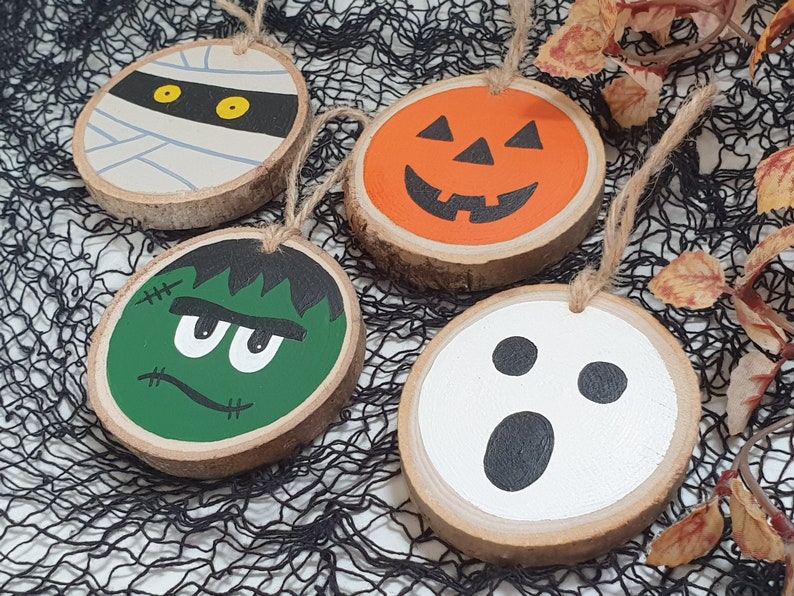 May include: Four wooden ornaments with Halloween themes. A mummy, a pumpkin, a Frankenstein, and a ghost. Each ornament is painted with a different character and has a string for hanging.