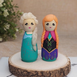 May include: Two hand-painted wooden dolls dressed as Elsa and Anna from the film Frozen. The dolls are standing on a wooden base. Elsa is wearing a blue and silver dress with a white braid. Anna is wearing a purple and green dress with a red braid.