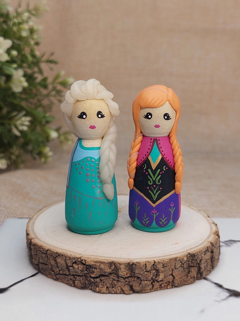 May include: Two hand-painted wooden dolls dressed as Elsa and Anna from the film Frozen. The dolls are standing on a wooden base. Elsa is wearing a blue and silver dress with a white braid. Anna is wearing a purple and green dress with a red braid.