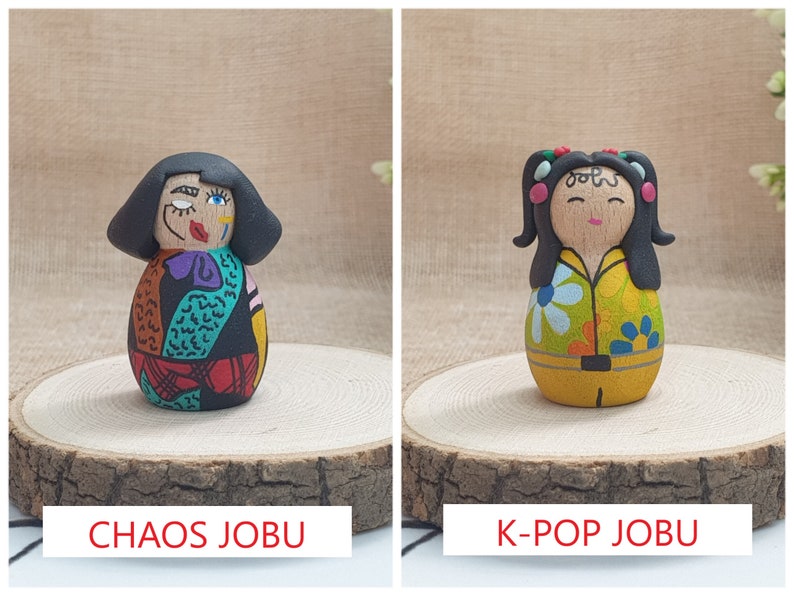 May include: Two hand-painted wooden dolls with black hair. The doll on the left has a colorful, abstract design. The doll on the right has a yellow shirt with a floral pattern and a black belt. The dolls are labeled "Chaos Jobu" and "K-Pop Jobu".
