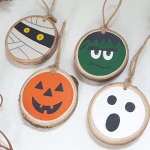May include: Four wooden ornaments with Halloween themes. The ornaments are painted with a mummy, a Frankenstein, a pumpkin, and a ghost. Each ornament has a twine loop for hanging.