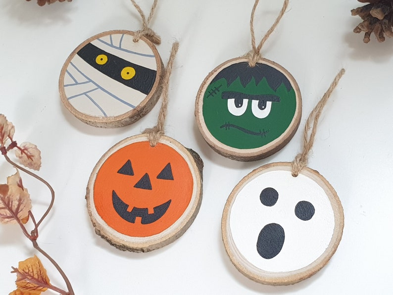 May include: Four wooden ornaments with Halloween themes. The ornaments are painted with a mummy, a Frankenstein, a pumpkin, and a ghost. Each ornament has a twine loop for hanging.