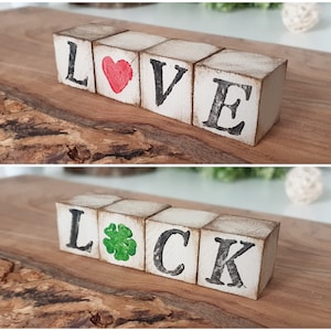 May include: Distressed wooden blocks spelling "LOVE" with a red heart and "LUCK" with a green shamrock. The blocks have black lettering and a vintage appearance. They are displayed on a wooden surface.
