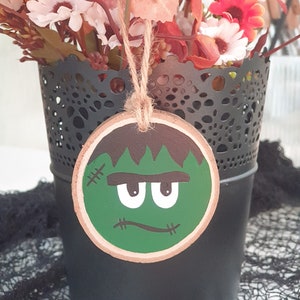 May include: A green and brown wooden ornament with a cartoon Frankenstein face. The ornament is hanging from a string and is in front of a black vase with a lace pattern.