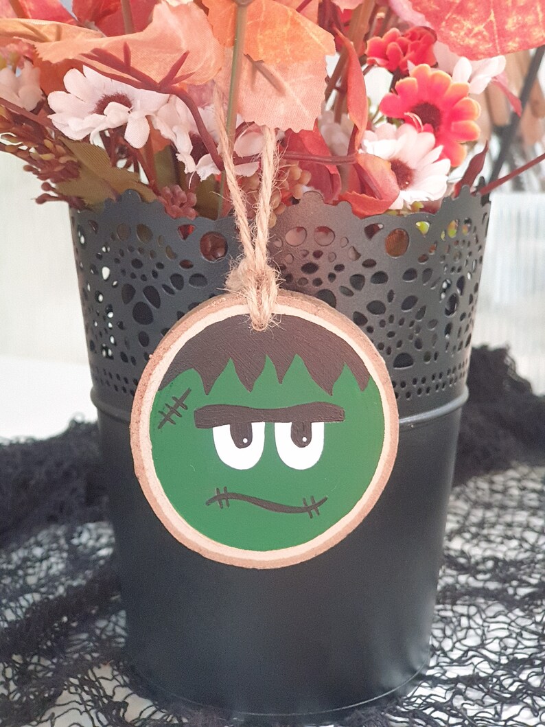 May include: A green and brown wooden ornament with a cartoon Frankenstein face. The ornament is hanging from a string and is in front of a black vase with a lace pattern.