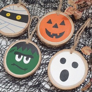 May include: Four Halloween-themed wooden ornaments with painted faces. The ornaments are a mummy, a pumpkin, a Frankenstein monster, and a ghost.