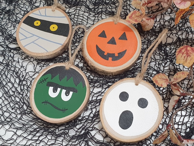 May include: Four Halloween-themed wooden ornaments with painted faces. The ornaments are a mummy, a pumpkin, a Frankenstein monster, and a ghost.