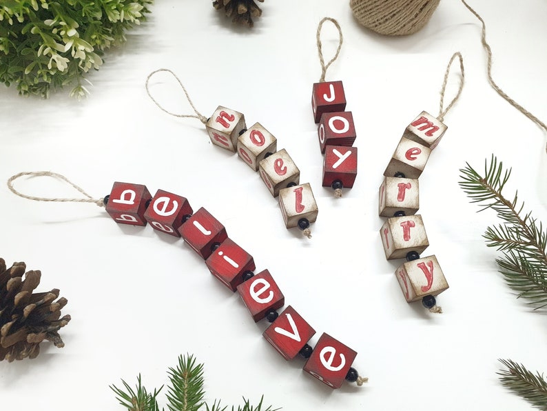 May include: Four red wooden ornaments with white lettering spelling out "believe", "joy", "merry", and "home". Each ornament is strung together with twine and black beads.