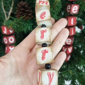May include: A wooden Christmas decoration spelling out "Merry" in red letters on white blocks. The blocks are strung together with black beads and twine.