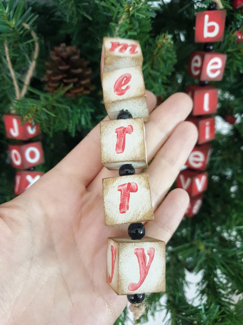 May include: A wooden Christmas decoration spelling out "Merry" in red letters on white blocks. The blocks are strung together with black beads and twine.