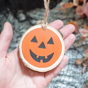 May include: A wooden pumpkin ornament with a painted orange face and black eyes, nose, and mouth. The ornament has a string attached for hanging.