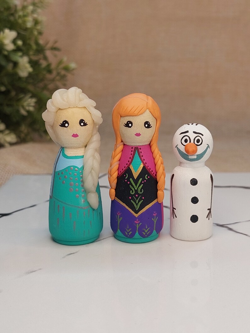 May include: Three hand-painted wooden peg dolls, depicting characters from the film Frozen. The dolls are Elsa, Anna, and Olaf. Elsa has a blue dress and blonde hair, Anna has a purple dress and red hair, and Olaf is a snowman with a carrot nose.