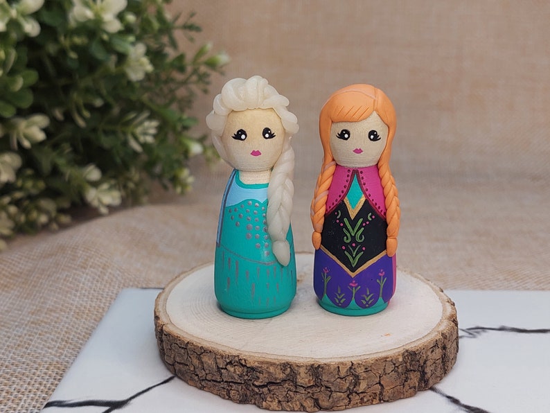 May include: Two handmade wooden peg dolls dressed as Elsa and Anna from the movie Frozen. Elsa is wearing a blue and silver dress with a white braid. Anna is wearing a purple and pink dress with a red braid.