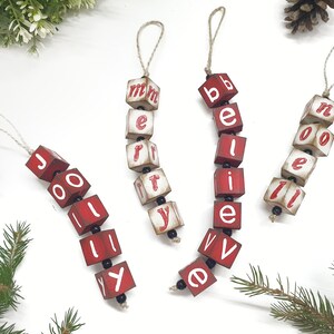 May include: Five red wooden ornaments with white letters spelling out "Jolly", "Merry", "Believe", and "Noel". The ornaments are strung together with twine and black beads.