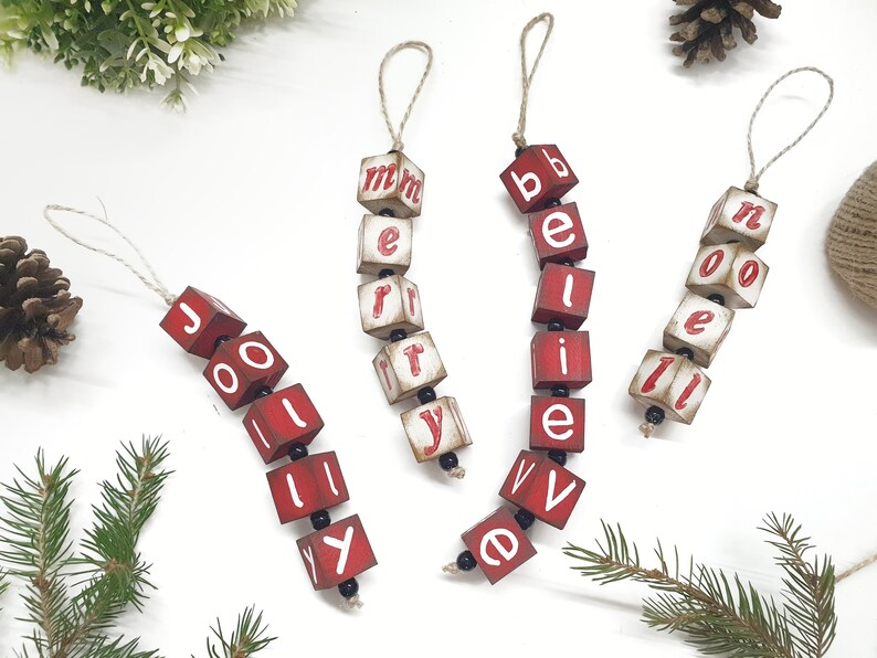 May include: Five red wooden ornaments with white letters spelling out "Jolly", "Merry", "Believe", and "Noel". The ornaments are strung together with twine and black beads.
