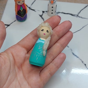 May include: A hand-painted wooden doll of Elsa from Frozen, wearing a teal dress with silver glitter accents and a white braid. The doll has a painted face with pink cheeks and blue eyes.