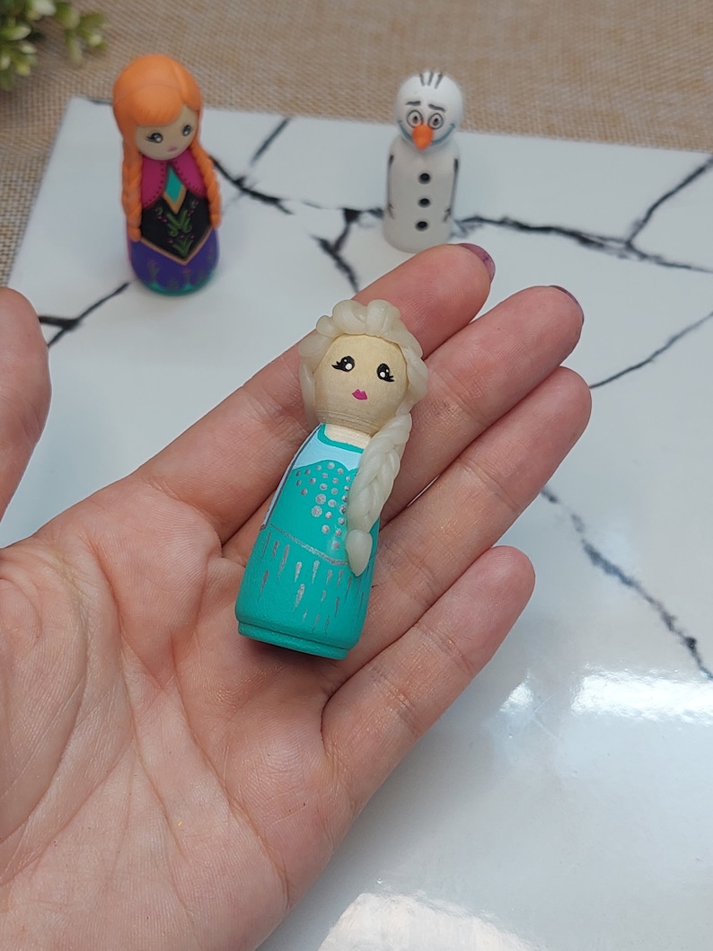May include: A hand-painted wooden doll of Elsa from Frozen, wearing a teal dress with silver glitter accents and a white braid. The doll has a painted face with pink cheeks and blue eyes.